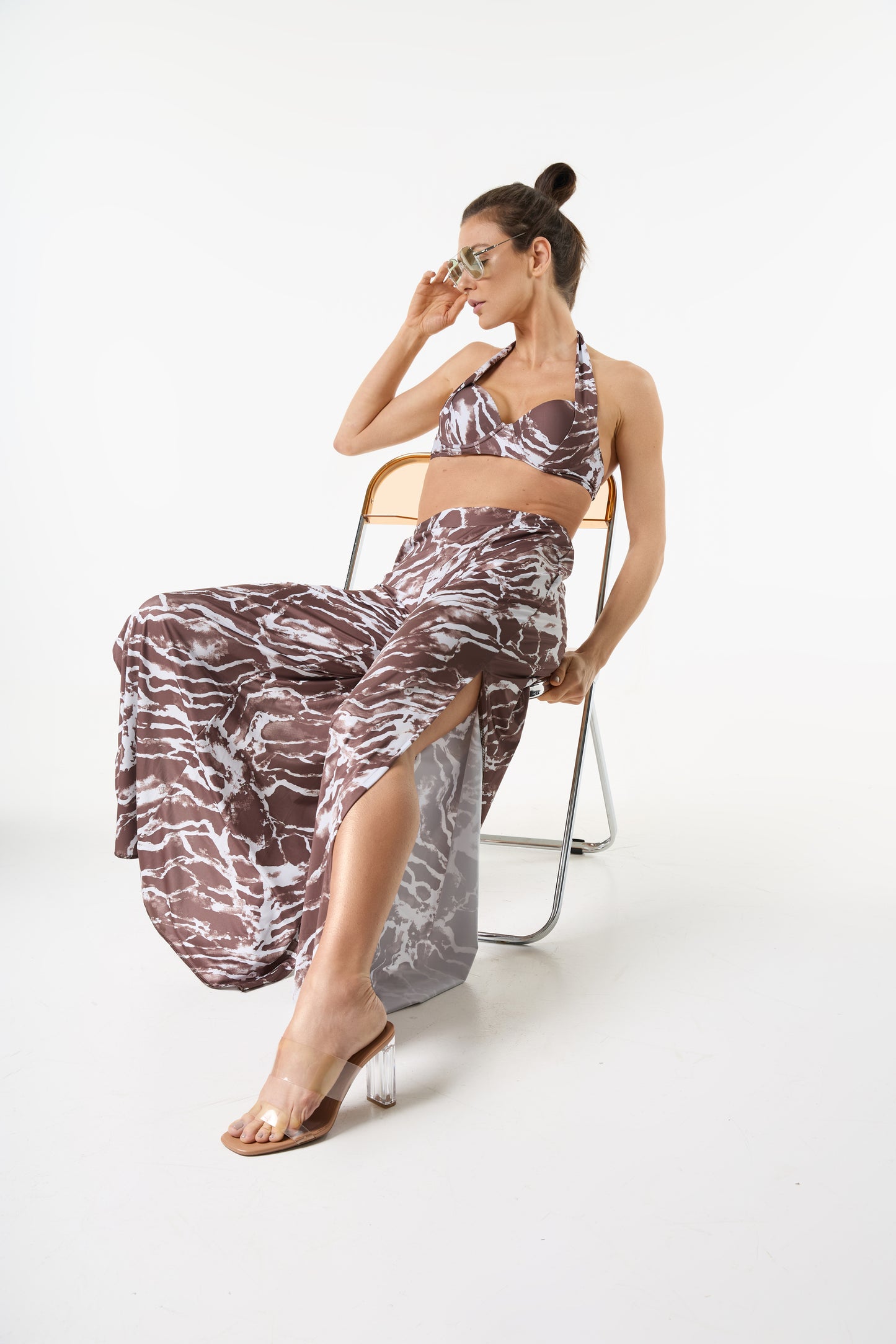 Conjunto top e calça com estampa marmorizada marrom e branca. A modelo está sentada de lado em uma cadeira transparente, ajustando os óculos, com as pernas cruz