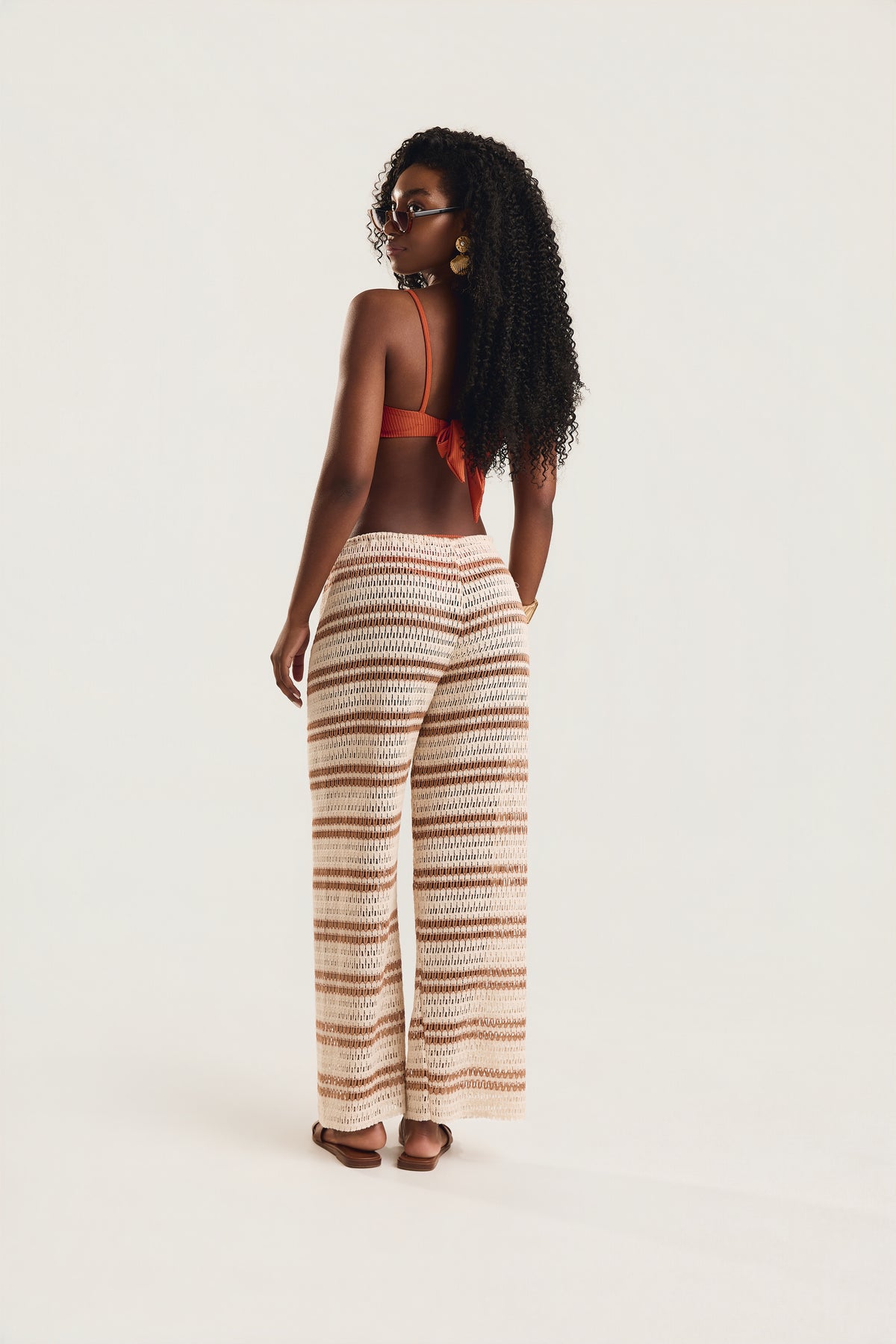 Calça pantalona de crochê listrada em off white caramelo, modelo de costas em postura ereta com olhar sobre o ombro