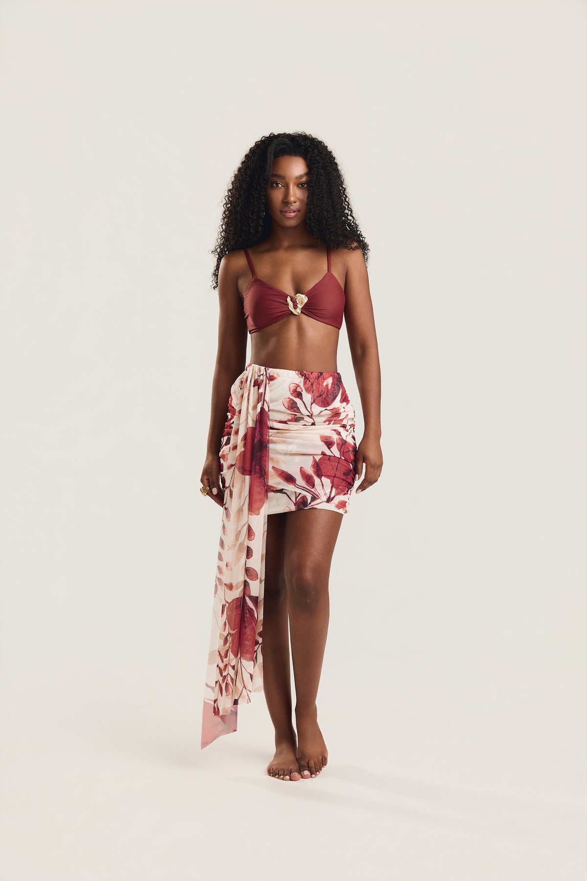 Modelo de frente, postura ereta com braços relaxados ao lado do corpo e olhar direto para a câmera, usando saia mini franzida com estampa floral vermelho