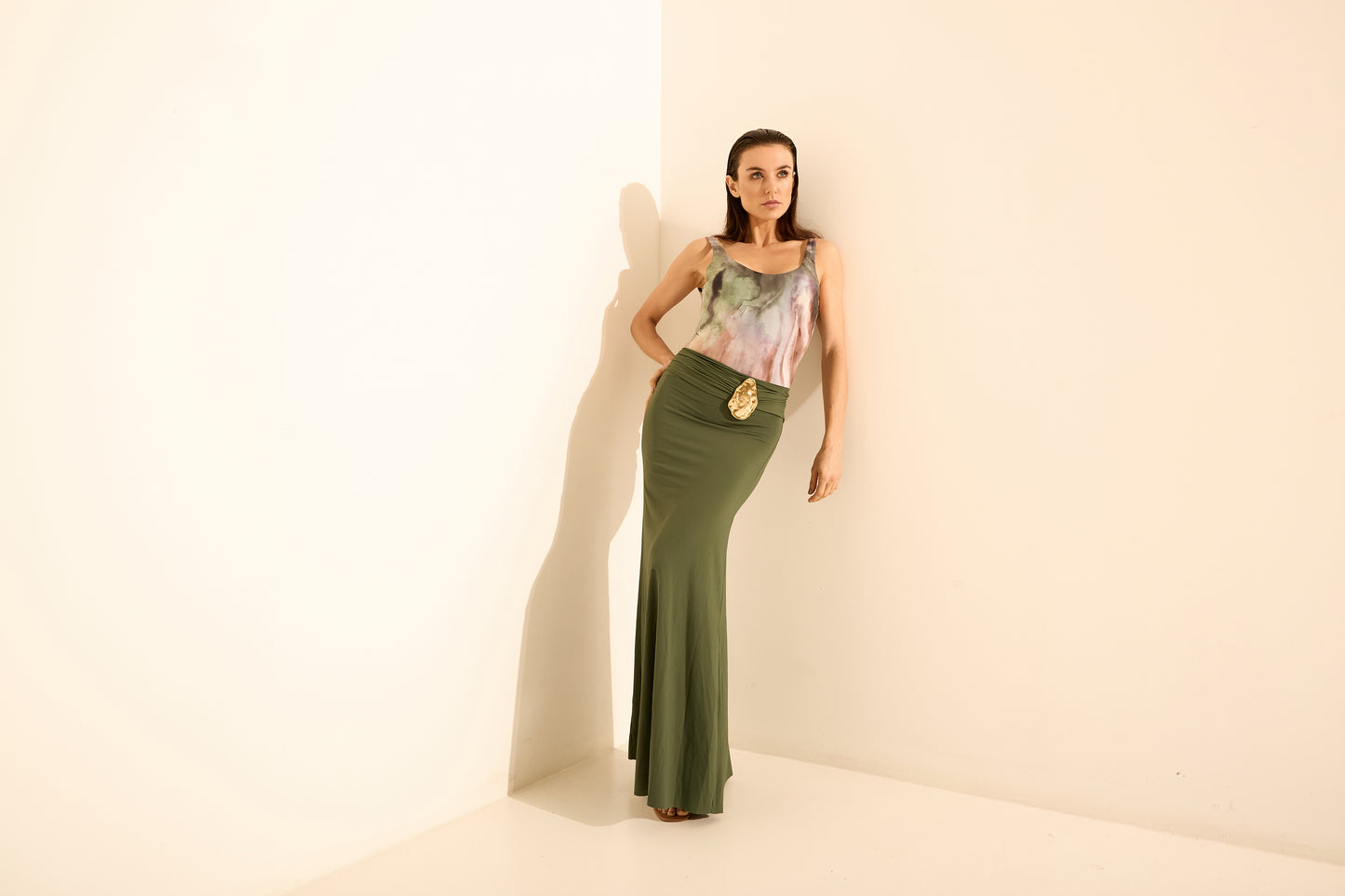 Look saia longa verde oliva blusa estampada. A modelo posa de frente em um canto, com a mão na cintura e a blusa com estampa aquarelada em tons past