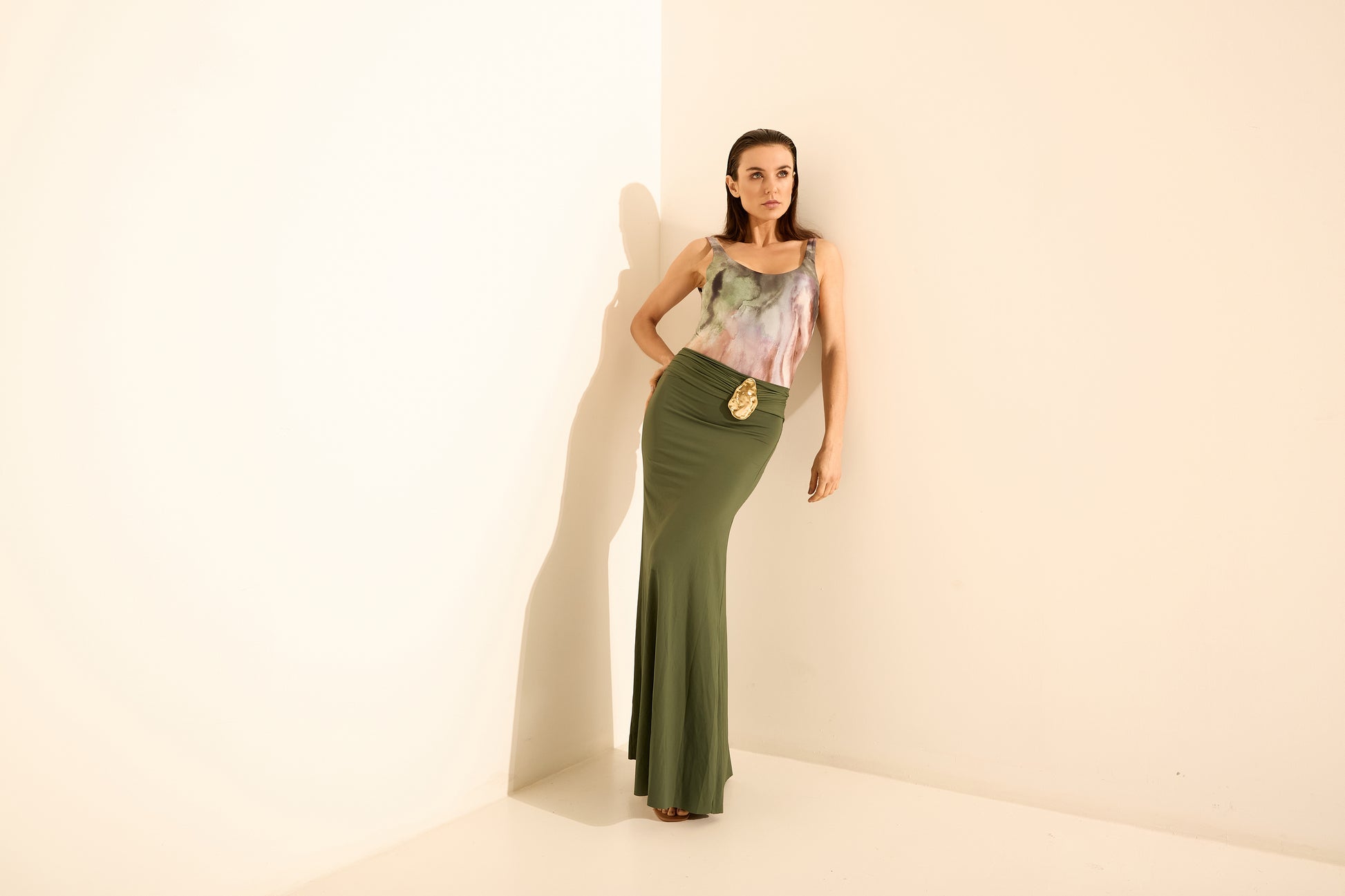 Look saia longa verde oliva blusa estampada. A modelo posa de frente em um canto, com a mão na cintura e a blusa com estampa aquarelada em tons past