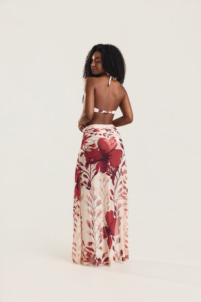 Modelo em pé de costas, olhando levemente para o lado, usando saia longa Mareblu com estampa floral vermelha em fundo off-white. Vista traseira evidencia o caimento fluido da saia.