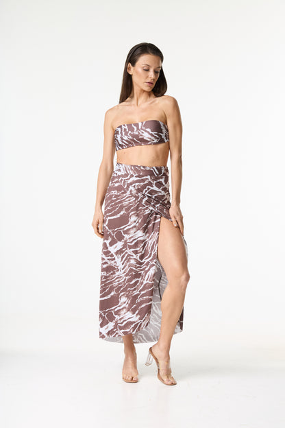 Conjunto de top faixa e saia longa com fenda, apresentando estampa abstrata em tons de marrom e branco. A modelo posa de frente, com a perna direita exposta pela fenda da saia.
