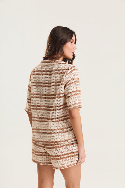 Modelo em pé de costas, levemente virada para o lado, olhar direcionado para longe da câmera, vestindo conjunto de crochê listrado off-white e caramelo, fundo neutro claro
