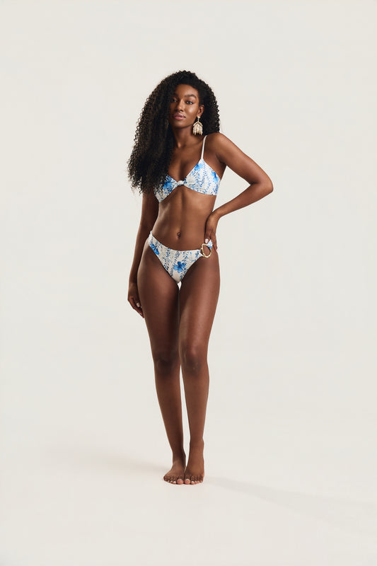Modelo em posição frontal com mão na cintura, usando biquíni top com nó e calcinha metal lateral em estampa floral azul e branco
