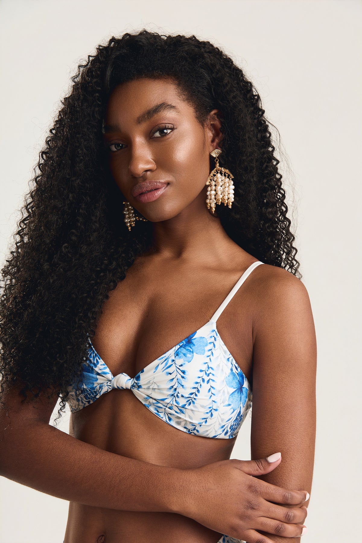 Close no rosto e busto da modelo com braços cruzados, usando top com nó em estampa floral azul e branco
