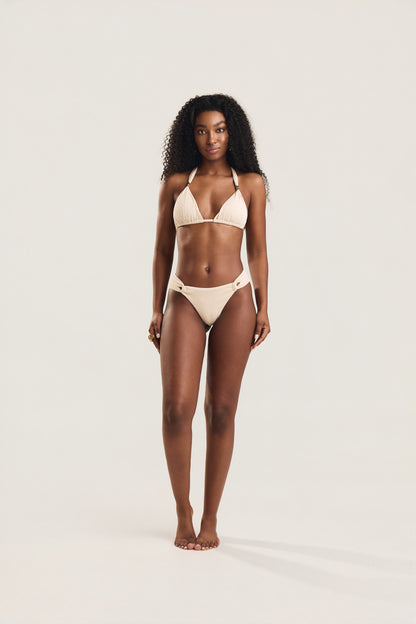 Modelo em posição frontal, corpo inteiro, usando biquíni Mareblu com top cortininha com bojo e detalhe em metal dourado e calcinha frente dupla na cor off-white textura