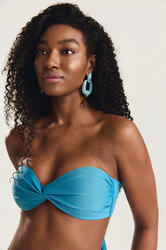 Close no rosto e busto da modelo, usando top tomara que caia azul textura com detalhe torcido frontal e brincos argola azul