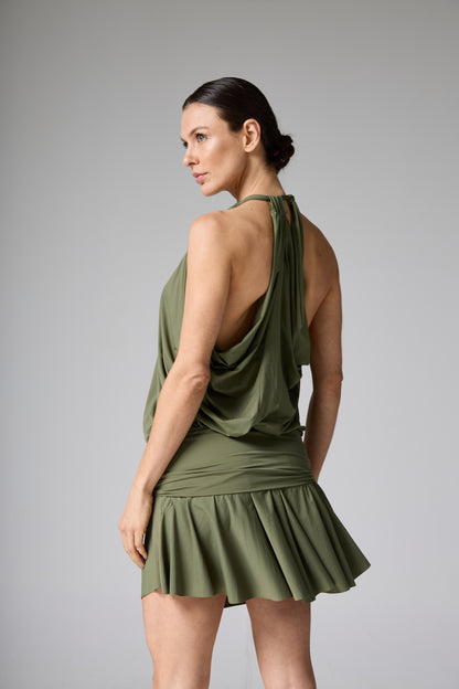 Vestido mini verde oliva com decote halter drapeado e saia plissada. A modelo está de costas, com a cabeça virada, destacando o caimento da parte superior e o movimento da saia.