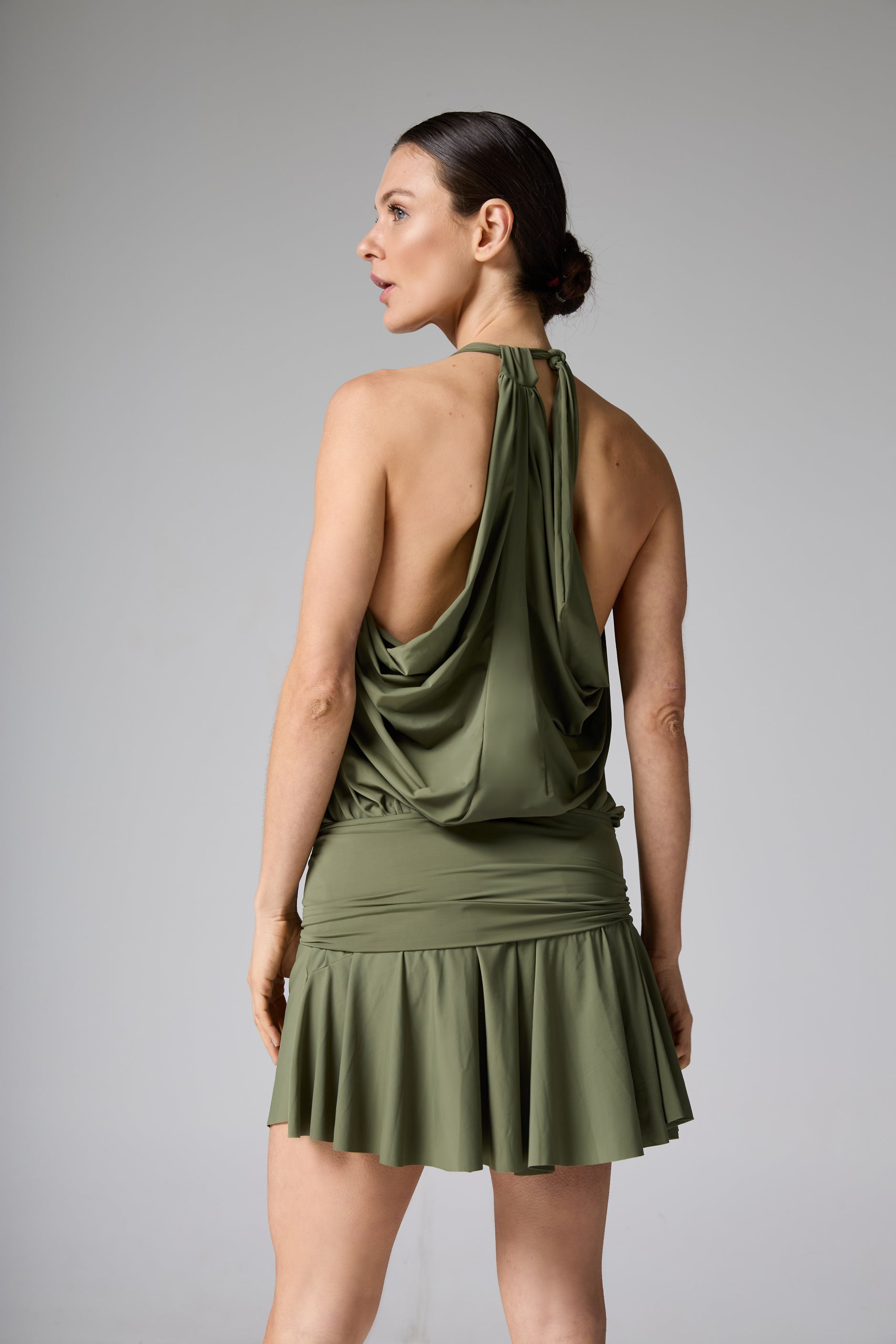 Maiô vestido verde-oliva. A modelo é vista de costas, com a cabeça virada, destacando o decote frente única e o drapeado aberto nas costas.