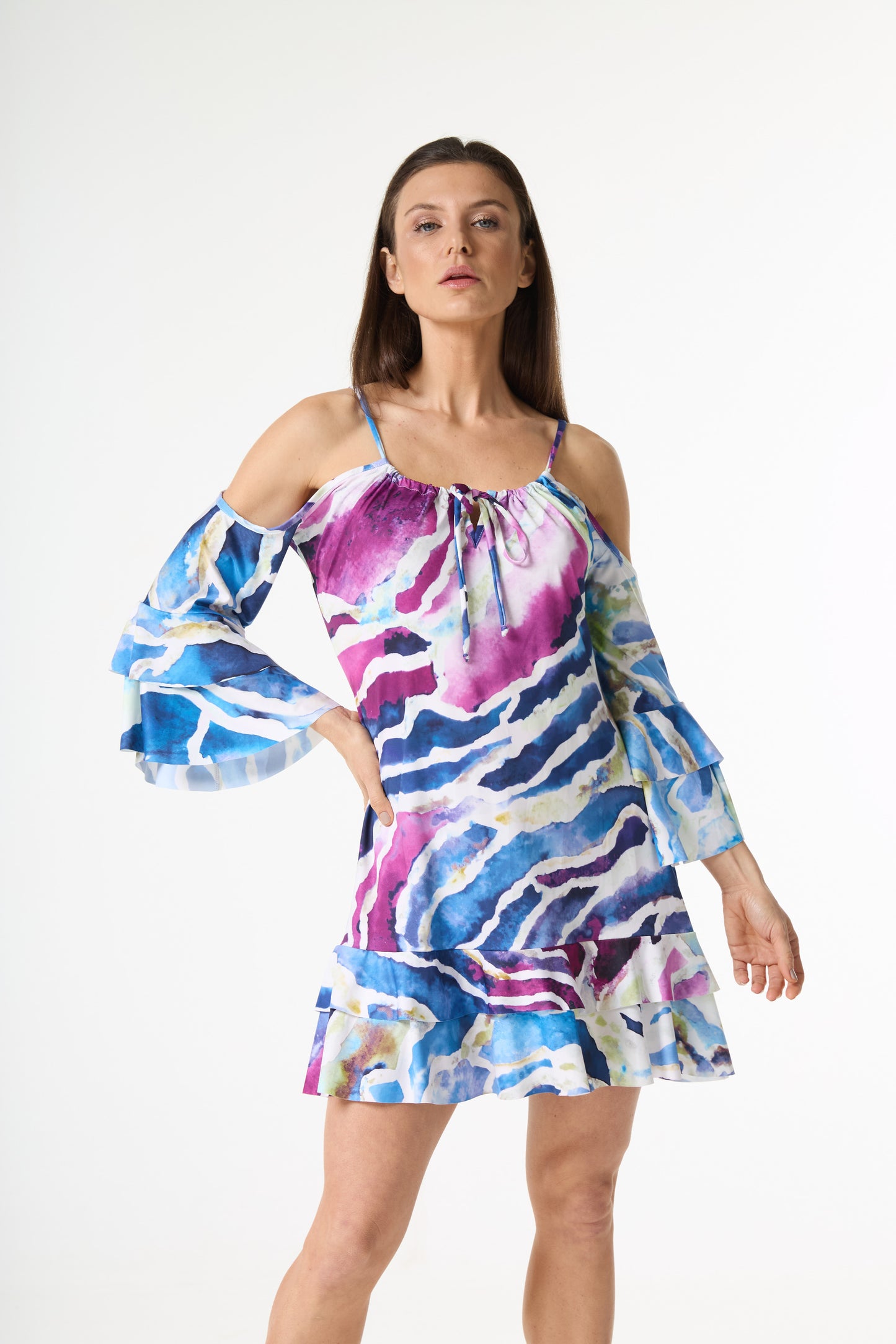 Vestido curto estampa aquarela com alças finas, ombros à mostra e mangas longas com babados. A estampa abstrata em tons de azul, roxo e branco se destaca. A modelo posa de frente, com a mão esquerda na cintura, em fundo branco.