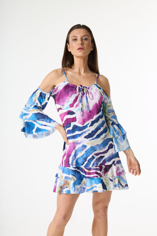 Vestido curto estampa aquarela com alças finas, ombros à mostra e mangas longas com babados. A estampa abstrata em tons de azul, roxo e branco se destaca. A modelo posa de frente, com a mão esquerda na cintura, em fundo branco.