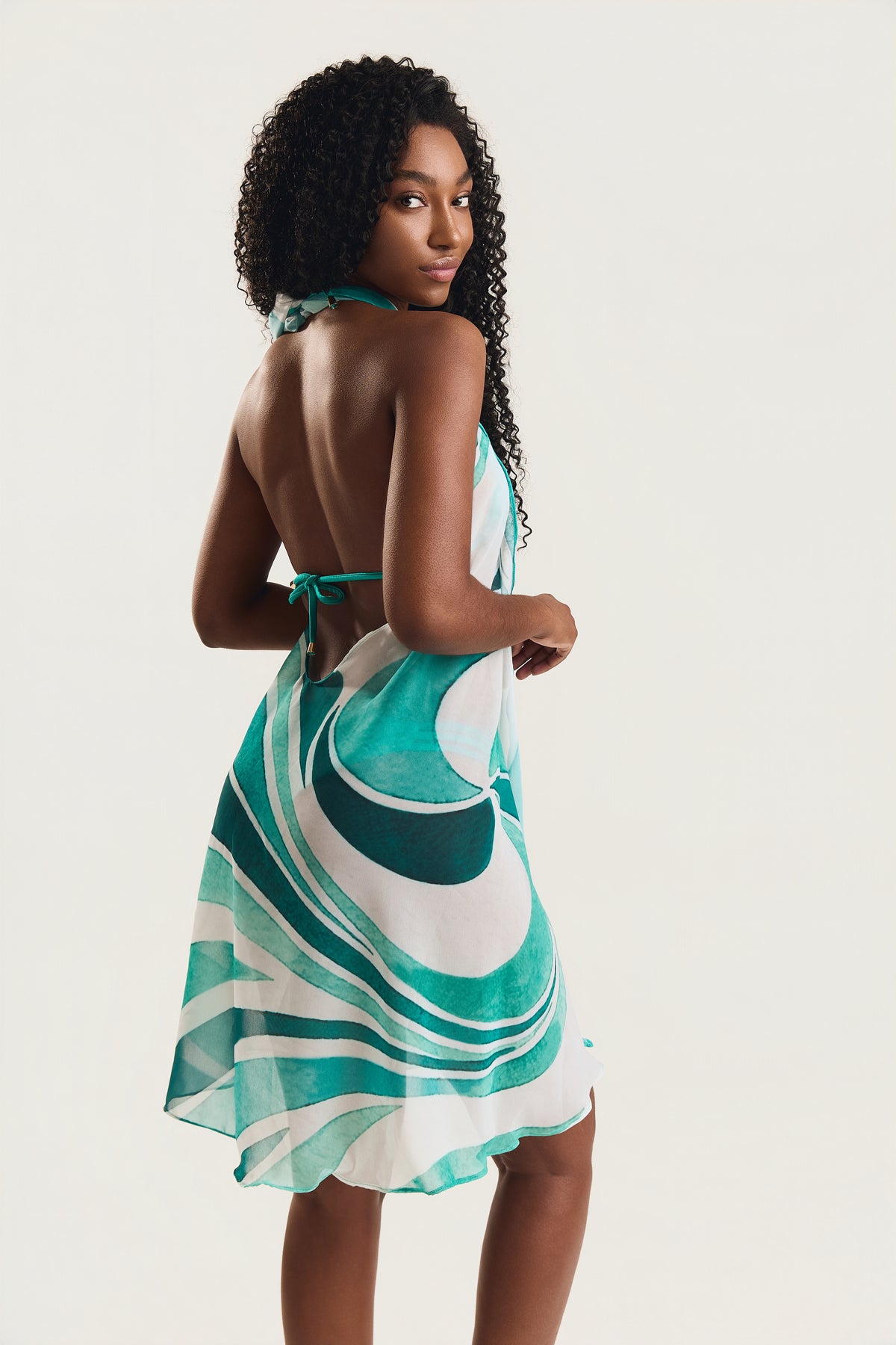 Vestido pareô estampado em geo verde, modelo de costas olhando por cima do ombro, decote aberto nas costas com amarração, fundo neutro