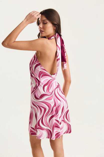 Vestido pareô estampado em geo rosa, modelo de costas em posição de três quartos com braço levantado à cabeça, decote aberto nas costas com amarração halter, fundo neutro