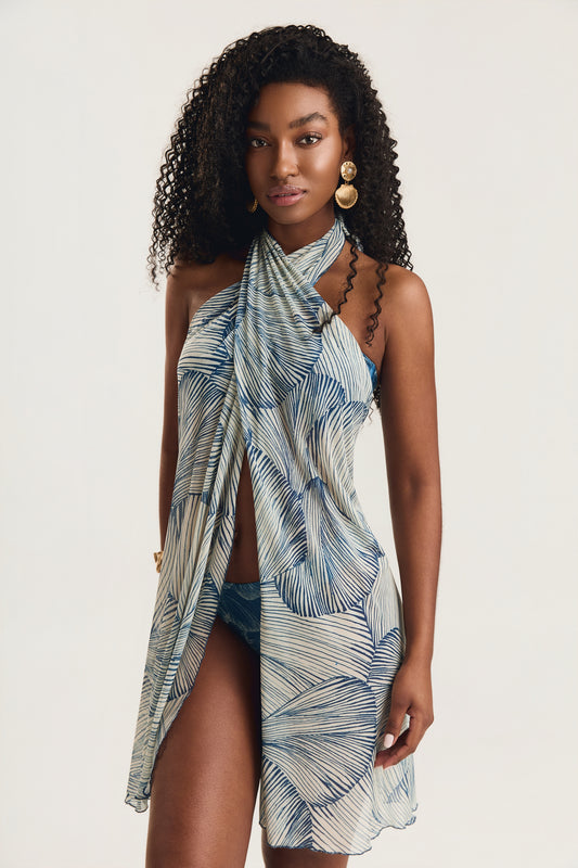 Vestido pareô com estampa de conchas azuis, modelo de frente em posição frontal com olhar direto para a câmera, decote halter cruzado, fundo neutro