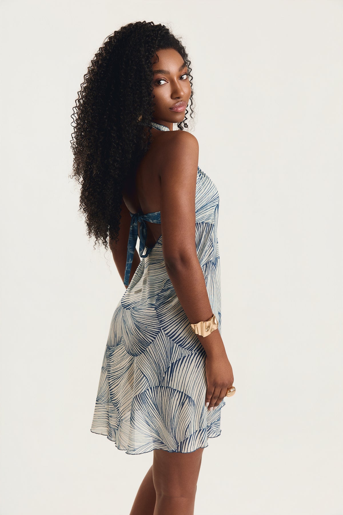 Vestido pareô com estampa de conchas azuis, modelo de costas em posição de três quartos olhando por cima do ombro, decote aberto nas costas, fundo neutro