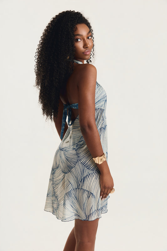 Vestido pareô com estampa de conchas azuis, modelo de costas em posição de três quartos olhando por cima do ombro, decote aberto nas costas, fundo neutro