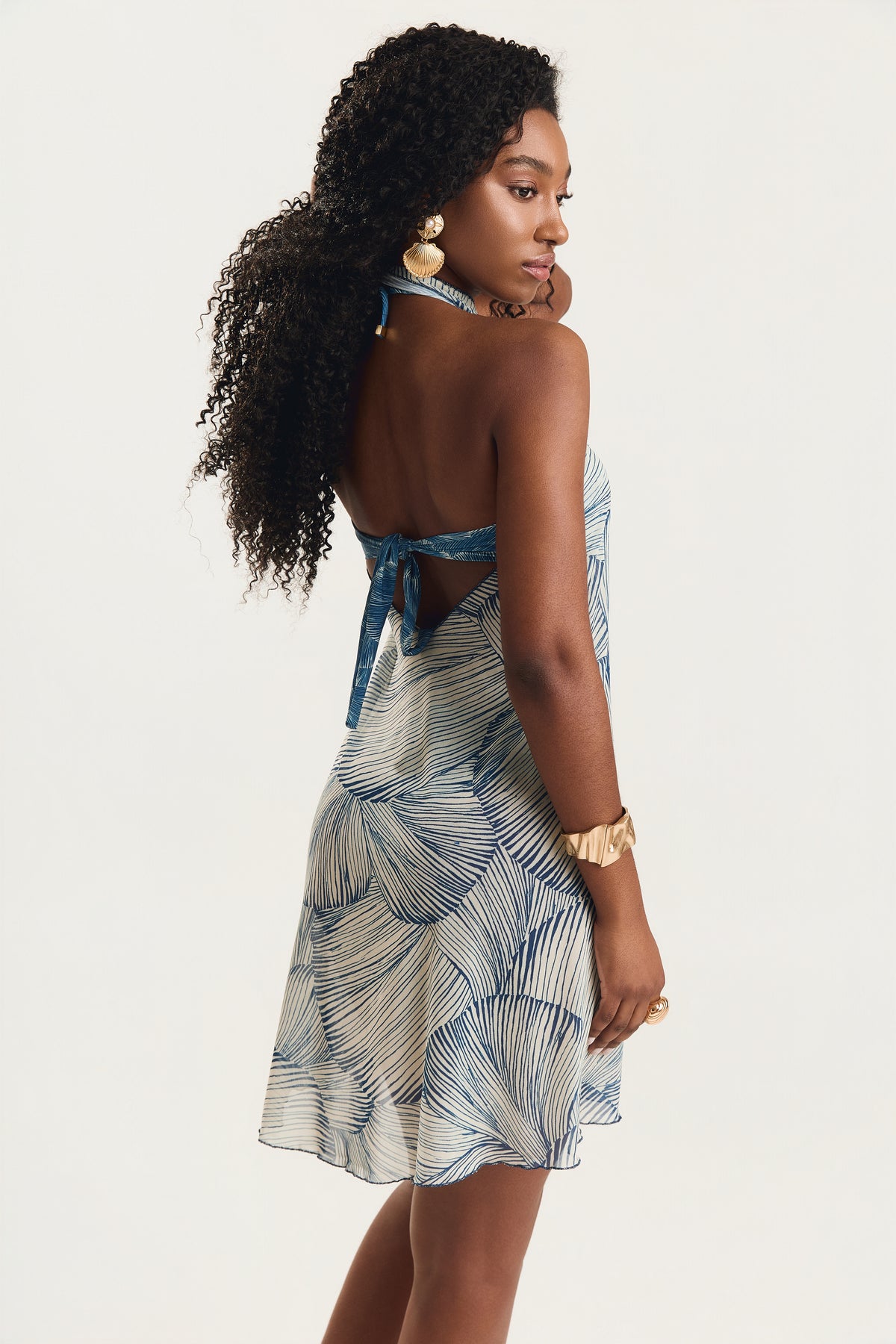 Vestido pareô com estampa de conchas azuis, modelo de costas em posição lateral com olhar para baixo, decote aberto nas costas, fundo neutro