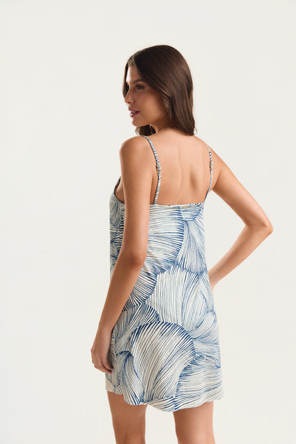  Modelo em pé de costas com olhar voltado sobre o ombro direito, corpo inteiro, usando o Vestido de Alcinha Franzido Mareblu com estampa Concha Azul evidenciando as alças finas e o decote nas costas