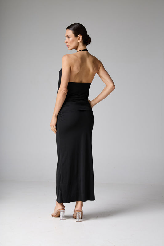 Vestido longo preto. A modelo está de costas, levemente virada, com uma das mãos na cintura, destacando o decote halter, as costas nuas e o caimento da sa