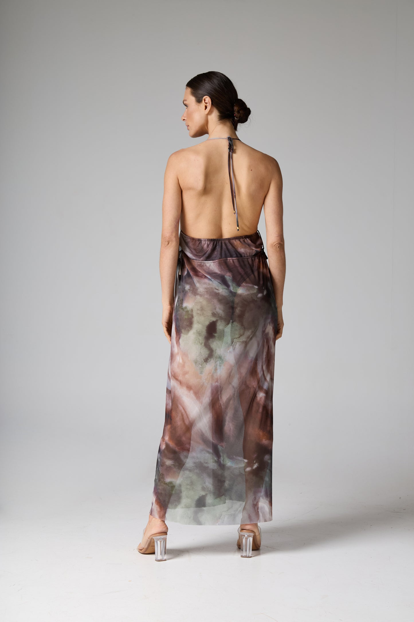 Vestido longo estampado costas nuas com padrão abstrato em tons terrosos, como marrom e verde. A modelo está de costas, mostrando o caimento solto e transparente do tecido, e as alças finas amarradas no pescoço.